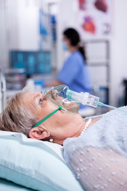 Máscara de oxígeno que ayuda a una anciana a respirar mientras está acostada en la cama del hospital debido a una infección por coronavirus