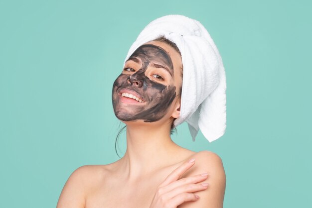 Máscara de spa de mujer, retrato saludable del concepto de belleza. Mascarilla facial de barro, mascarilla facial de arcilla spa. Hermosa mujer con procedimiento facial de barro cosmético, concepto de salud spa. Tratamiento de belleza para el cuidado de la piel. Toalla en la cabeza.