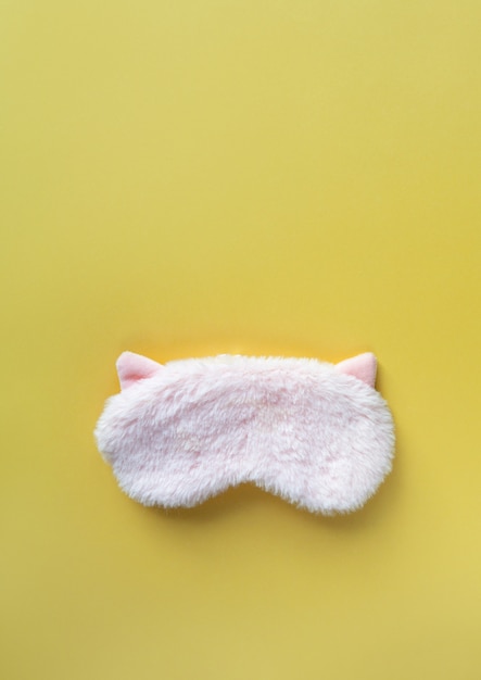 Máscara de sueño de piel esponjosa rosa pastel con orejas pequeñas sobre fondo de papel amarillo pastel. Vista superior, plano, copia espacio. Concepto de sueños vívidos. Accesorios para niñas y mujeres jóvenes.