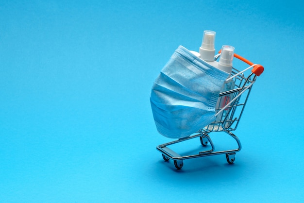 Máscaras protectoras y gel desinfectante desinfectante antibacteriano en un pequeño carrito de la compra sobre fondo azul con espacio de copia.
