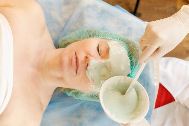 Mascarilla de colágeno. Tratamiento de la piel facial. Mujer recibiendo procedimiento cosmético en salón de belleza. Botella con crema hidratante en el fondo.