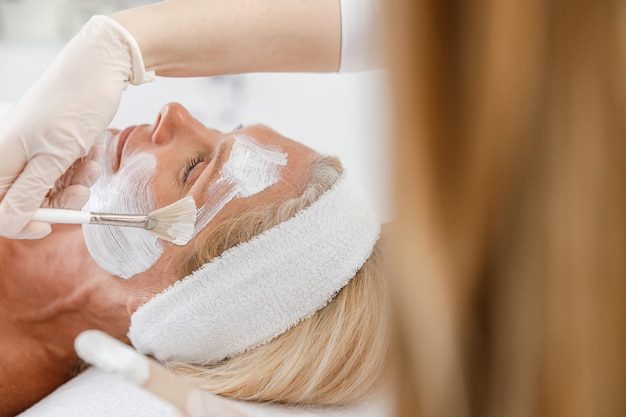 Mascarilla exfoliante facial de cerca, tratamiento de belleza spa, cuidado de la piel. Mujer mayor recibiendo cuidado facial por esteticista en el salón de spa.