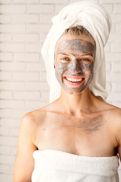 Mascarilla Facial Spa. Spa y belleza. Mujer sonriente joven vistiendo toallas de baño blancas con una mascarilla facial de arcilla en su rostro