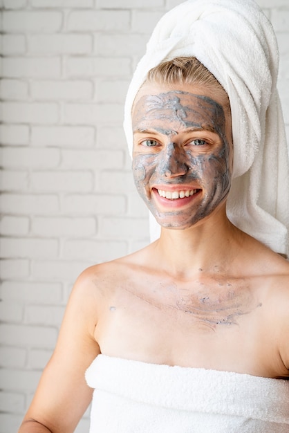 Mascarilla Facial Spa. Spa y belleza. Mujer sonriente joven vistiendo toallas de baño blancas con una mascarilla facial de arcilla en su rostro