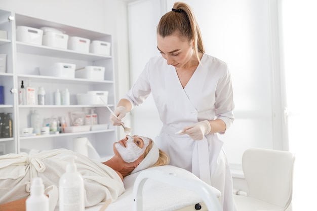 Mascarilla, tratamiento de belleza spa, cuidado de la piel. Mujer mayor recibiendo cuidado facial por esteticista en el salón de spa, vista lateral.