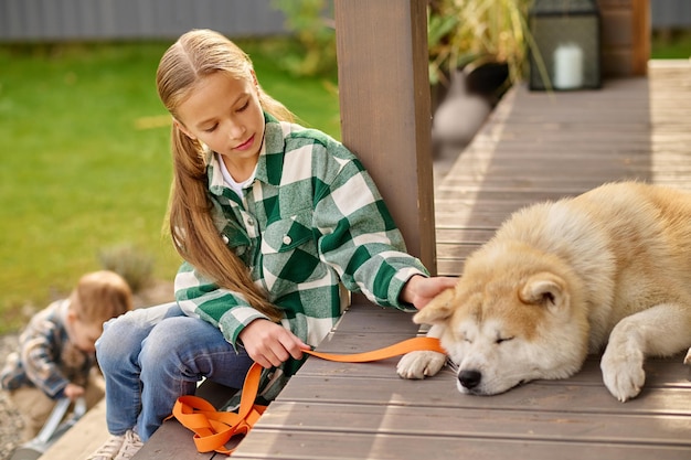 Mascota. Linda niña en edad escolar con cabello largo y rubio en camisa a cuadros y jeans sentado en el porche viendo tocar perro dormido acostado con correa y hermano pequeño detrás