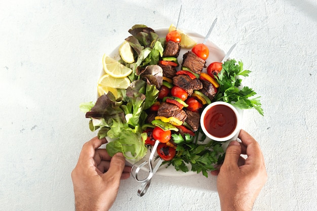 Masculinas mãos segurando um prato com kebab de carne grelhada com legumes e molho vista superior