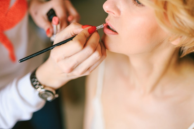 Maskenbildnerin in einem weißen Hemd malt die Mädchenlippen mit roter Lippenstiftnahaufnahme