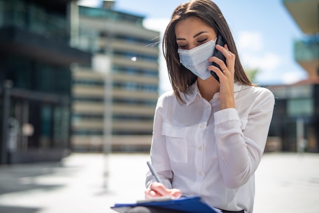 Maskierte frau, die am telefon spricht, während sie notizen schreibt covid und coronavirus-konzept