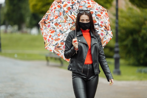 maskiertes Mädchen geht die Straße entlang. Mädchen in einer Schutzmaske geht im Park mit einem Regenschirm im Regen. Coronavirus-Infektion COVID-19