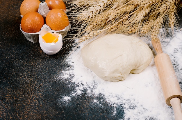 Massa de fermento crua em uma tigela e ingredientes, ovos, farinha, espigas de trigo na mesa escura, conceito de padaria