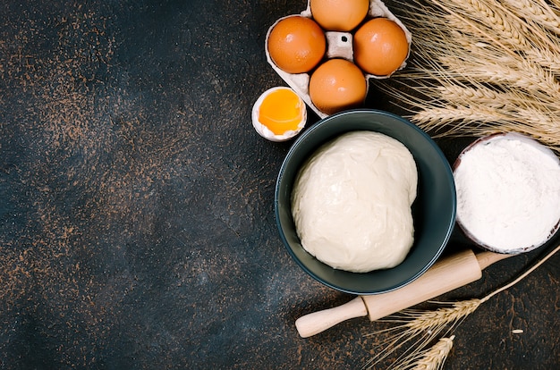 Massa de fermento crua na tigela e ingredientes, ovos, farinha, espigas de trigo no escuro