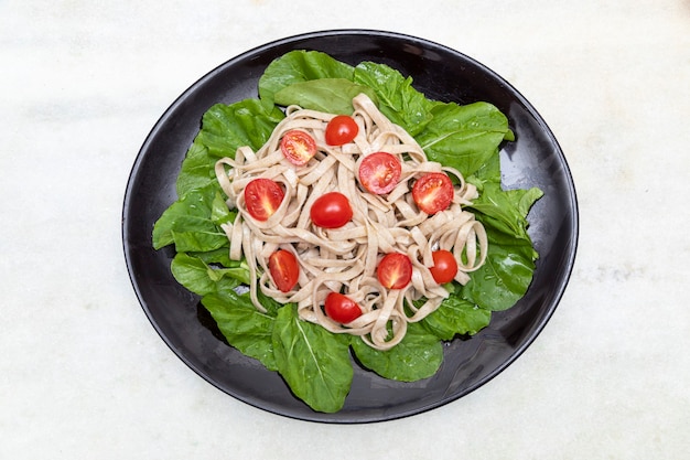 Massa integral com folhas verdes de rúcula e tomate cereja em placa preta e fundo de mármore branco