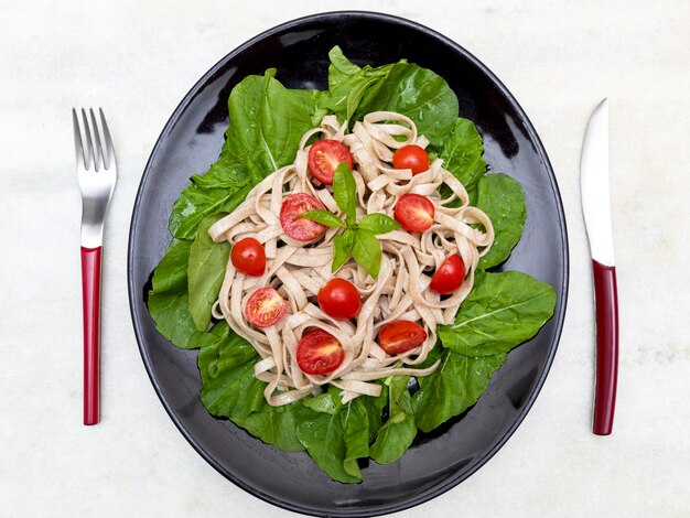 Massa integral com folhas verdes de rúcula e tomate cereja em placa preta e fundo de mármore branco