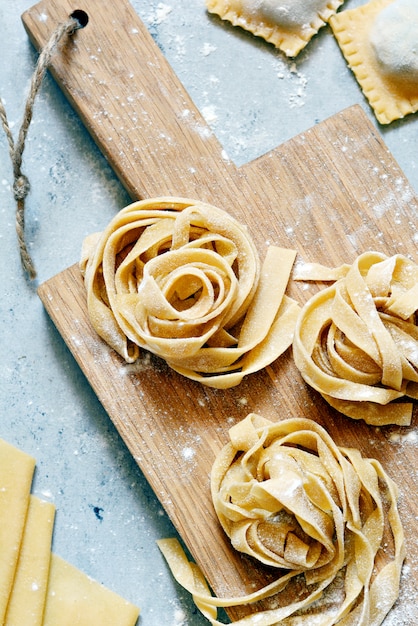 Massa italiana caseira, ravioli, fettuccine, tagliatelle em uma placa de madeira e em um fundo azul. o processo de cozimento, macarrão cru