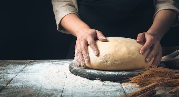 Massa nas mãos do cozinheiro em uma placa de madeira entre farinha em um fundo escuro.