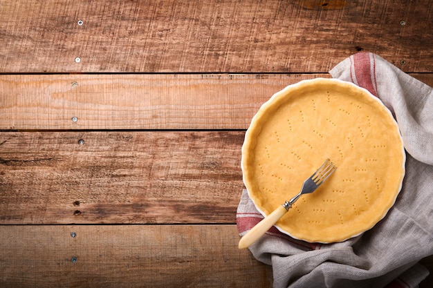 Massa para assar quiche, torta ou torta em forma de cozimento de cerâmica pronta para assar na toalha de cozinha sobre fundo de prancha rústica de madeira velha. Vista superior, copie o espaço. Cozimento caseiro de conceito para as férias.