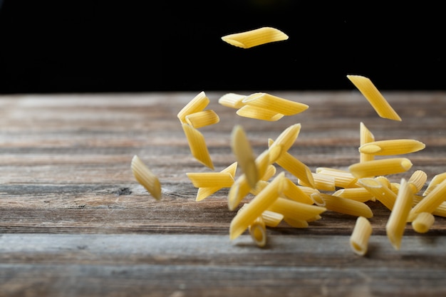 Massa penne caindo. Macarrão cru amarelo voando sobre fundo preto e rebote da mesa de madeira. DOF raso.