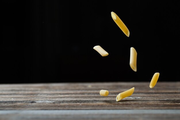 Massa penne caindo. Macarrão cru amarelo voando sobre fundo preto e rebote da mesa de madeira. DOF raso.