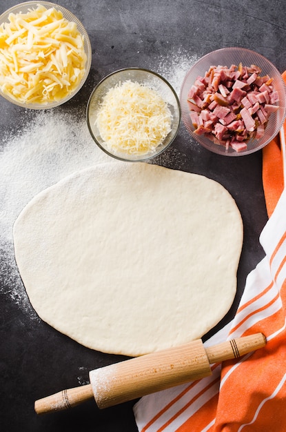 Massa rolada, farinha, enchimento da pizza e pino do rolo em um quadro-negro escuro.