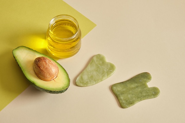 Massage mit Avocadoöl-Konzept Avocadoöl in einem Glas in Scheiben geschnittene Avocados und Gua-Sha-Scheiben auf grünem Hintergrund