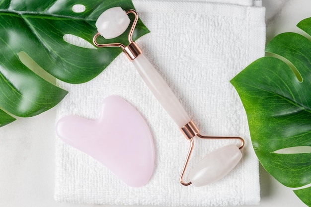 Massageador Gua Sha e rolo de rosto em fundo de toalha branca com folhas de monstera Ferramentas de massagem para cuidados com a pele facial conceito de tratamento de beleza SPA