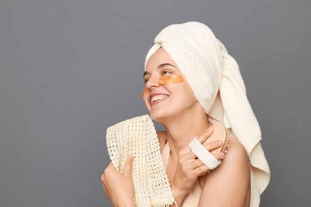 Massagem a seco Procedimentos de beleza no chuveiro Encantada jovem sorridente enrolada na toalha com manchas sob os olhos fazendo procedimentos de cuidados com a pele posando isolado sobre fundo cinza