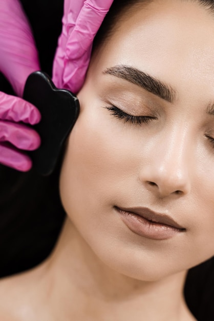 Massagem de guache para rosto de meninas usando raspador de pedra closeup Esteticista faz massagem Gua Sha para melhorar a circulação sanguínea e tonificar a pele facial