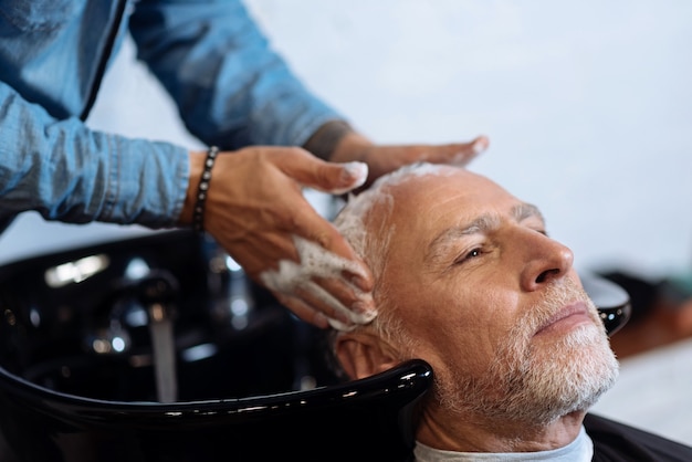 Massagem perfeita. Retrato do close-up de um homem barbudo agradável, tendo seu cabelo lavado por cabeleireiro na barbearia.