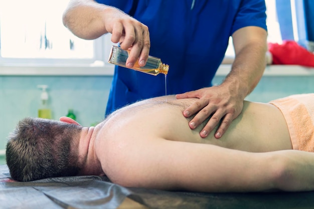 Massagista de massagem esportiva usando óleo de massagem paciente do sexo masculino em uma consulta com um fisioterapeuta para um procedimento de massagem