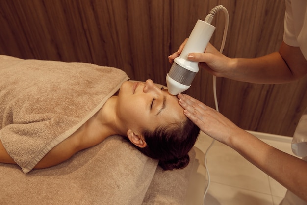 Massagista feminina com massageador vibratório faz massagem facial para mulher jovem. Massagem e relaxamento, cuidados com o corpo e a pele. Mulher atraente em salão de spa