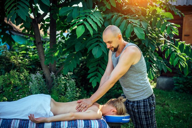 Massagista masculino dando bem-estar volta massagem ao ar livre. Mulher caucasiana, deitado na cama de massagem no jardim verde do centro de spa. Jovem mulher saudável desfrutando relaxante massagem corporal