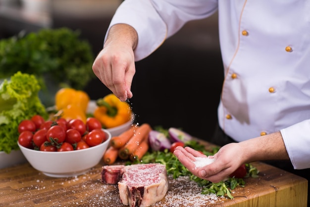 Master Chef pone sal en una jugosa rebanada de bistec crudo con verduras alrededor de una mesa de madera