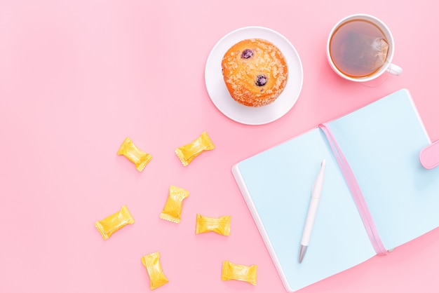 Material de escritório de mesa de trabalho, chá quente, doces e bolo em fundo rosa pastel