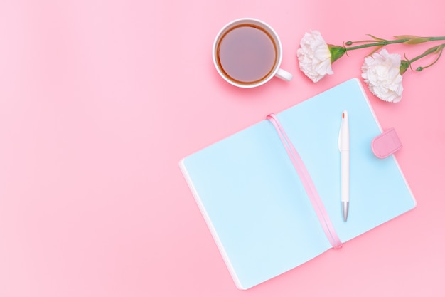 Material de oficina del escritorio del área de trabajo, té caliente y flor blanca sobre fondo rosa pastel