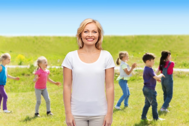 maternidade, filhos, paternidade e conceito de pessoas - mulher sorridente em camiseta branca em branco sobre fundo de grupo de crianças ao ar livre