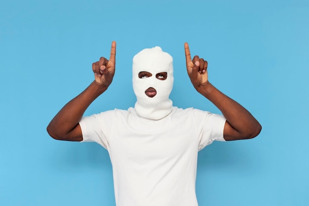 Foto matón afroamericano en balaclava blanca levanta las manos y apunta hacia arriba en el fondo azul aislado