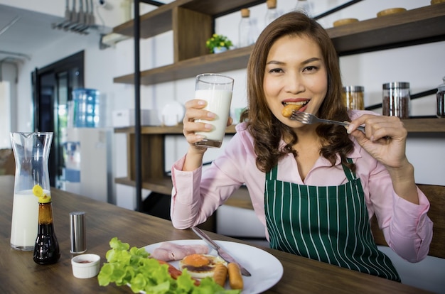 Foto matue erwachsene asiatische hausfrau in schürze, die am tisch in der küche sitzt und ein glas milch hält und bereit zum frühstück ist fröhliche süße frau, die morgens mit leckerem essen in den neuen tag startet