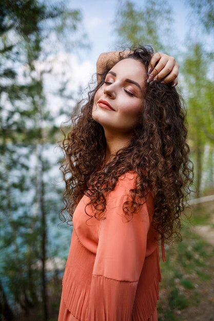 Mauerwerk aus Holz. Ein Mädchen mit langen lockigen Haaren in einem orangefarbenen Guipure-Kleid und Schuhen in der Natur, in einem Wald am See, stand in der Nähe von Bäumen und Büschen. Junge Frau lächelt und genießt das Leben