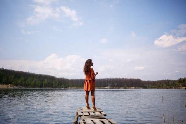 Mauerwerk aus Holz. Ein Mädchen mit langen lockigen Haaren in einem orangefarbenen Guipure-Kleid und Schuhen in der Natur, in einem Wald an einem See, stand in der Nähe von Bäumen und Büschen. Junge Frau lächelt und genießt das Leben