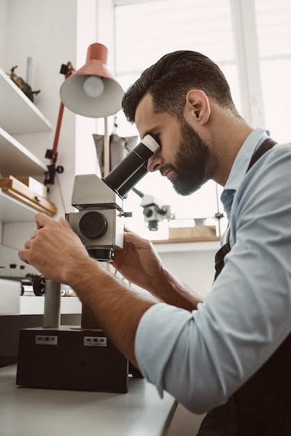 Máxima precisión. Vista lateral de un joyero masculino mirando el anillo a través del microscopio en un taller. Negocio. Equipo de joyería. Accesorios