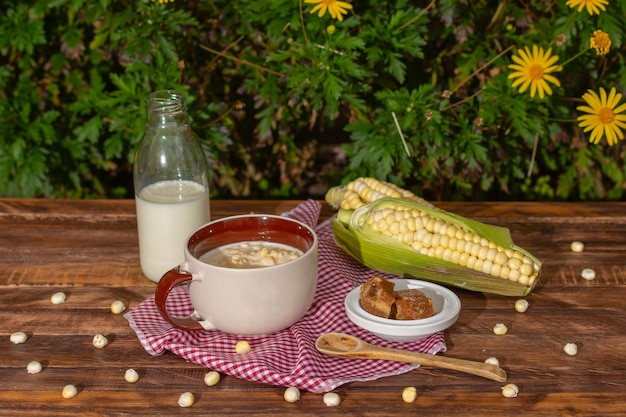 Mazamorra-Tasse mit Panela-Stücken, Milchflasche und Maiskolben über rustikalem Holztisch, breite gelbe Blumen im Hintergrund