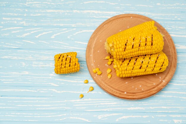 La mazorca de maíz caliente a la parrilla se encuentra en el fondo de la tabla de madera azul placa redonda para cortar. Copie el espacio para el texto.