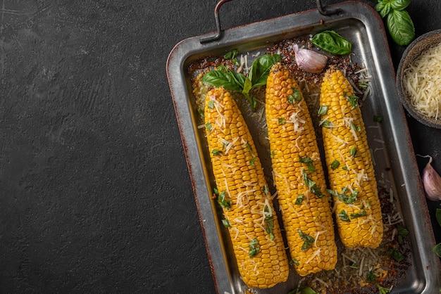 Mazorca de maíz a la plancha con mantequilla, queso parmesano y albahaca en una bandeja para hornear sobre fondo negro. vista superior con espacio de copia. comida vegetariana saludable