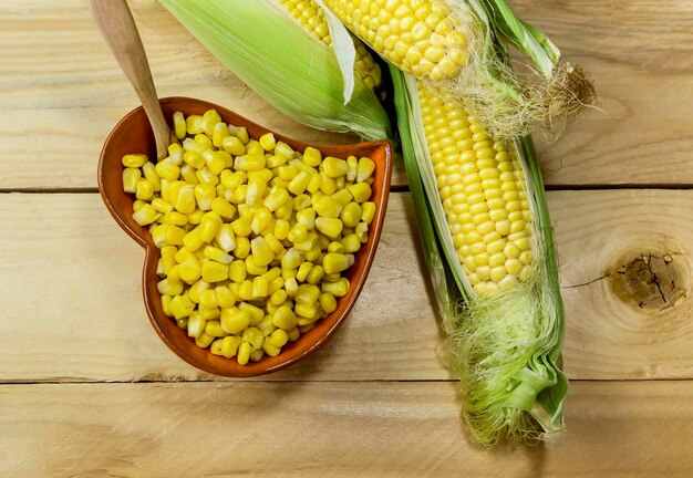 Las mazorcas de maíz crudas frescas y los granos de maíz hervidos en un tazón de cerámica en el primer plano de la mesa de madera