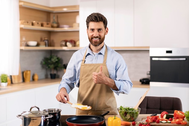 Me alegro del chef barbudo caucásico milenario en delantal preparando huevos revueltos y mostrando el pulgar hacia arriba