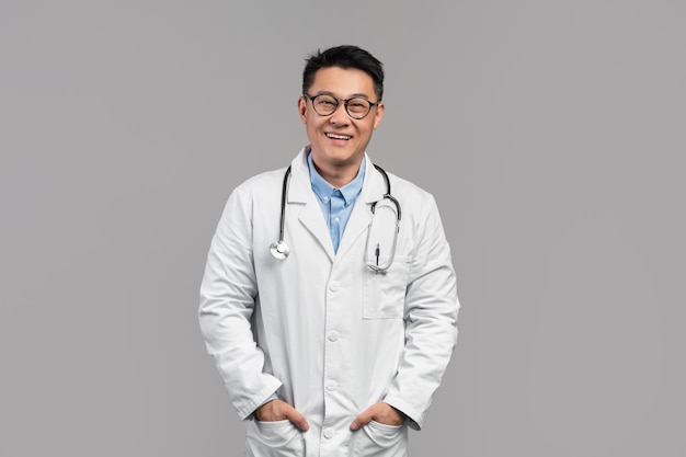 Me alegro de un médico japonés adulto atractivo y confiado con gafas de bata blanca con estetoscopio mirando a la cámara