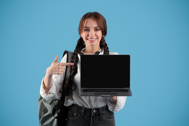 Me alegro de la mujer europea adolescente con coletas y mochila señalando con el dedo a la computadora portátil con pantalla en blanco