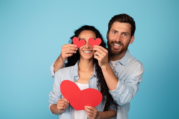 Me alegro de que el joven marido europeo con ropa informal ponga corazones en los ojos de la esposa árabe se divierta aislado de fondo azul