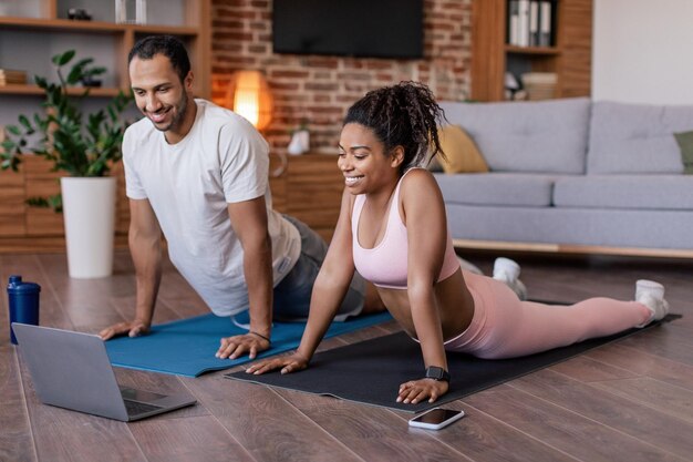 Me alegro de que una joven pareja negra con ropa deportiva se estire o practique yoga para ver una lección de video en una computadora portátil en una alfombra
